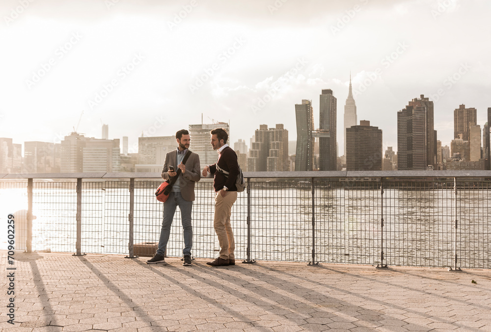 Young colleagues leaning on railings and talking by river at sunset in New York City