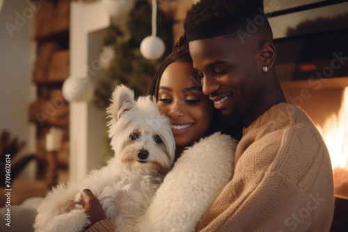 Beautiful African-American couple in white sweaters cuddles with a dog while sitting in front of the fireplace. photo