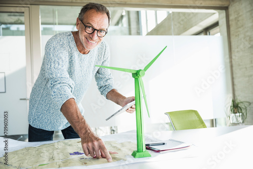 Smiling businessman pointing on blueprint and holding tablet PC near wind turbine model photo