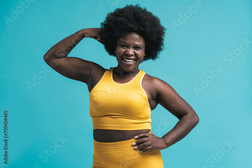 Cheerful young woman in yellow sports clothing flexing muscles against blue background photo
