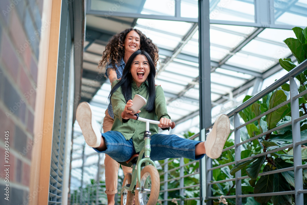 © Westend61 - Businesswoman enjoys riding bicycle with colleague pushing her in corridor
