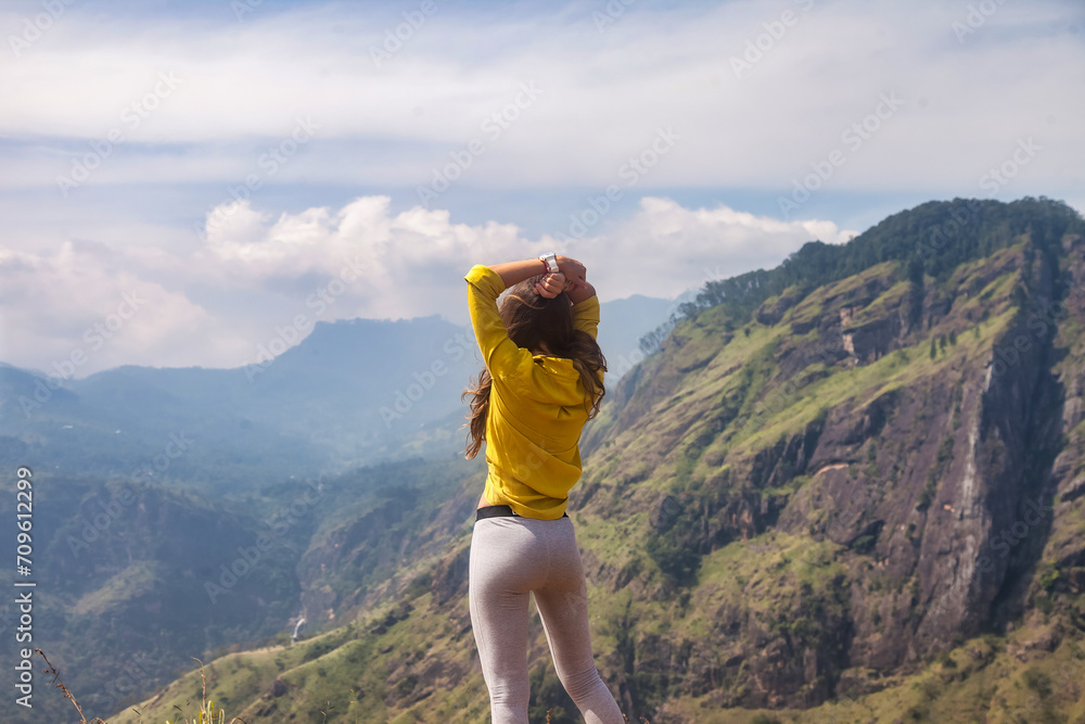 Rear view of adult woman enjoying at landscape Adam's peak mountain in tropical journey Sri Lanka, Ella. From behind of lady in yellow jacket posing at landmark nature background. Copy ad text space