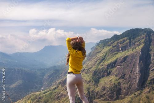 Rear view of adult woman enjoying at landscape Adam's peak mountain in tropical journey Sri Lanka, Ella. From behind of lady in yellow jacket posing at landmark nature background. Copy ad text space