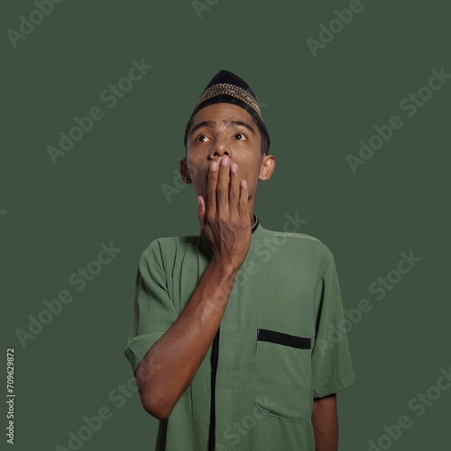 Oops Young Asian Muslim man covering mouth with hand looking at camera on green background