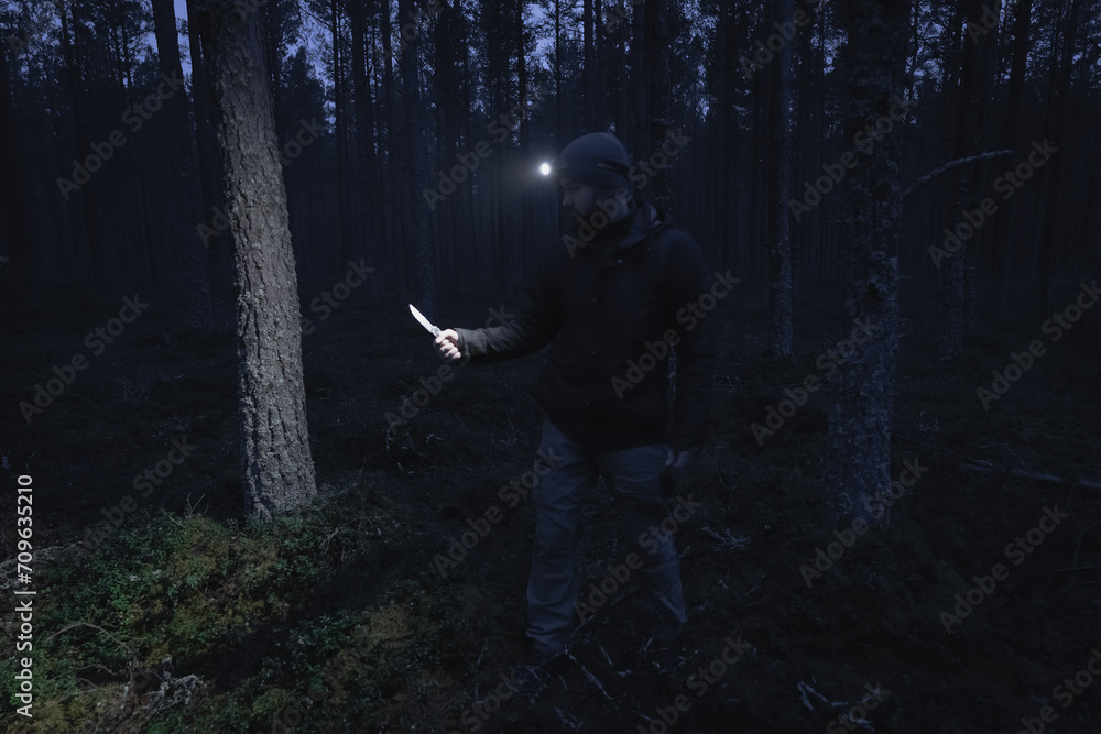 A man with a flashlight and a knife in his hand in a dark forest, photo with weak lighting.