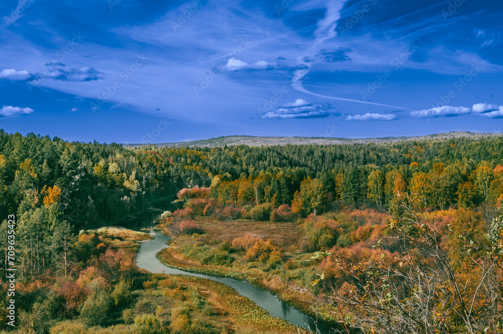 Obraz premium A winding river in the middle of an autumn forest with a blue sky with clouds.