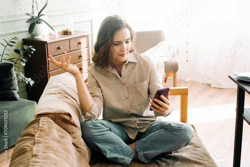 Digital Frustration. Perplexed and irate young woman experiences issues during a video call or online ordering in her living room. Technological Challenges and Emotional Distress photo