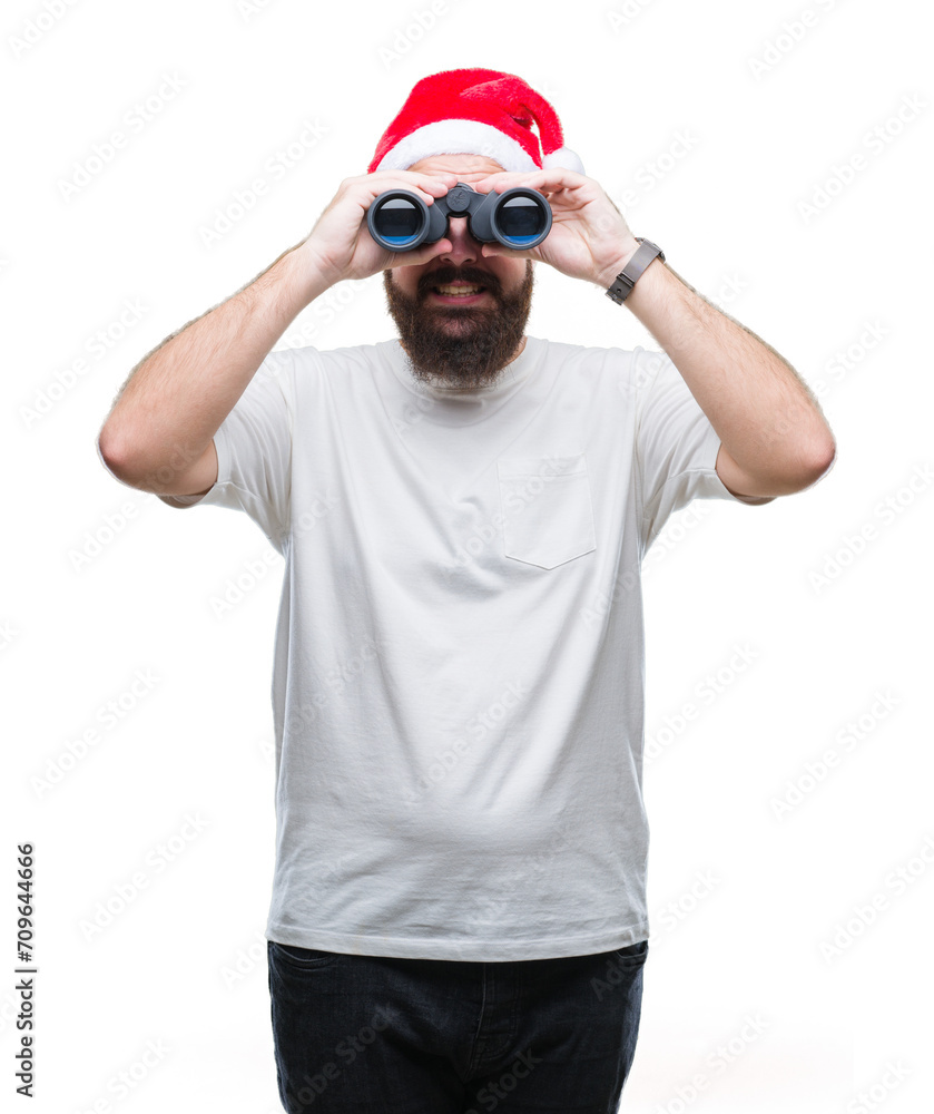 Young caucasian hipster man wearing christmas hat looking though binoculars over isolated background with a happy face standing and smiling with a confident smile showing teeth Young caucasian hipster man wearing christmas hat looking though binoculars over isolated background with a happy face standing and smiling with a confident smile showing teeth