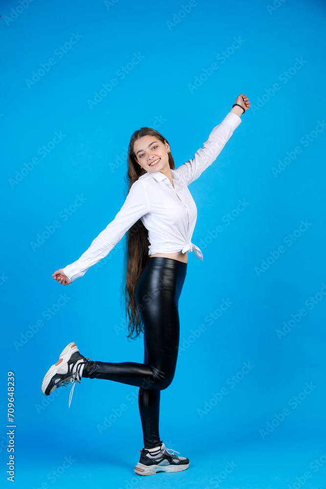 Obraz premium Carefree slender female raising hand, while having fun in studio. Side view of excited girl, bending leg, while posing to camera, isolated on blue background. Concept of fun.
