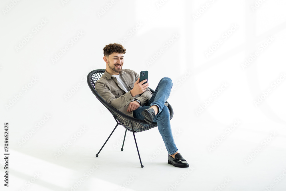 Portrait of casual cool mature man resting and using smart phone sitting on chair isolated on white studio background