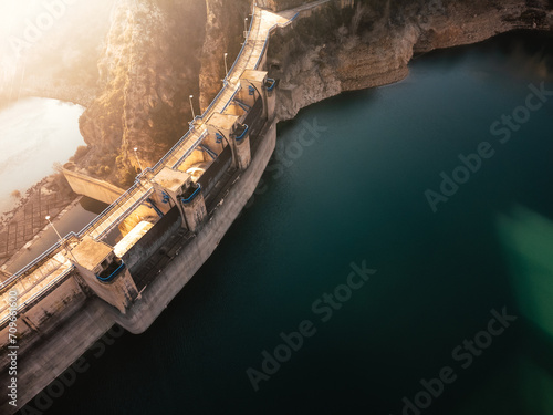 Sunlight bathes a dam with open spillways surrounded by steep cliffs above a deep blue reservoir