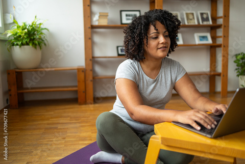 Fototapeta Naklejka Na Ścianę i Meble -  Young woman using laptop while sitting on yoga mat at home