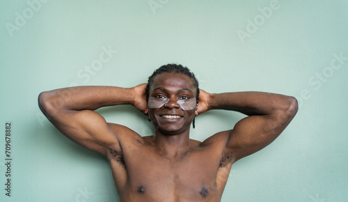 A joyful black man with under eye patches smiles at the camera hands behind his head against a mint green wall photo