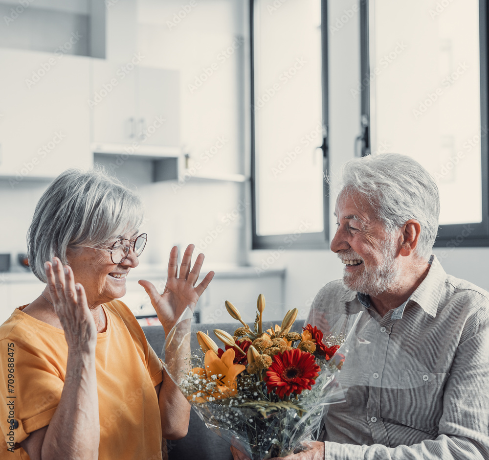 Old man giving flowers at his wife sitting on the sofa at home for the San Valentines’ day. Pensioners enjoying surprise together. In love people having fun.. Old man giving flowers at his wife sitting on the sofa at home for the San Valentines’ day. Pensioners enjoying surprise together. In love people having fun..