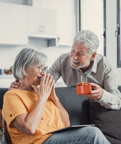 Smiling senior husband making surprise giving gift box to amazed wife, happy aged woman get unexpected present from spouse spending romantic weekend or celebrating anniversary together.