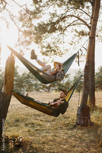 A guy and a girl are relaxing in hammocks in a pine forest at sunset. A couple in love rest in hammocks after a hike in the mountains, a guy plays the ukulele.
