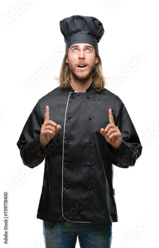 Young handsome cook man with long hair over isolated background amazed and surprised looking up and pointing with fingers and raised arms.
