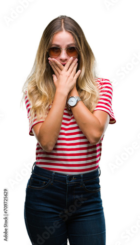 Young beautiful blonde woman wearing sunglasses over isolated background shocked covering mouth with hands for mistake. Secret concept.