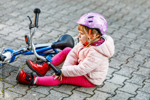 Cute little girl sitting on the ground after falling off her bike. Upset crying preschool child with safe helmet getting hurt while riding a bicycle. Active family leisure with kids.