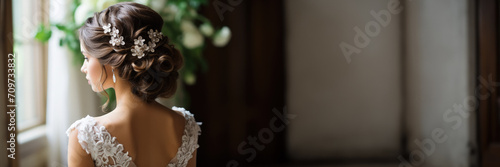 A bride in a lace wedding dress with an intricate hairstyle adorned with floral accessories gazes out a window in a soft, natural light setting