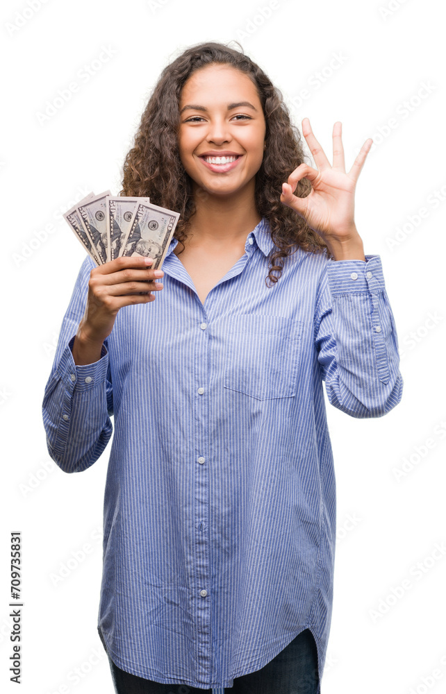 Young hispanic woman holding dollars doing ok sign with fingers, excellent symbol Young hispanic woman holding dollars doing ok sign with fingers, excellent symbol