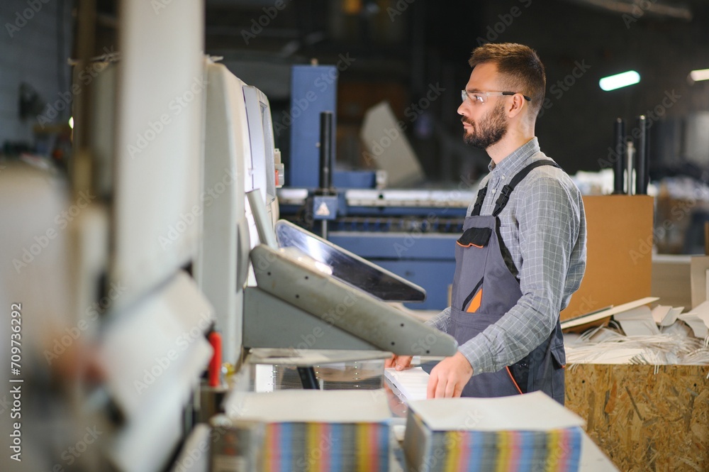 Printing house interior with modern offset print machine and operator in working uniform checking quality and controlling process of print