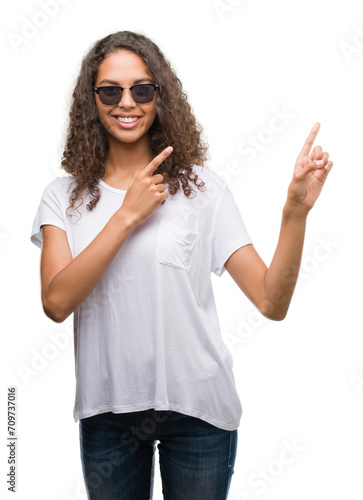 Young hispanic woman wearing sunglasses smiling and looking at the camera pointing with two hands and fingers to the side.