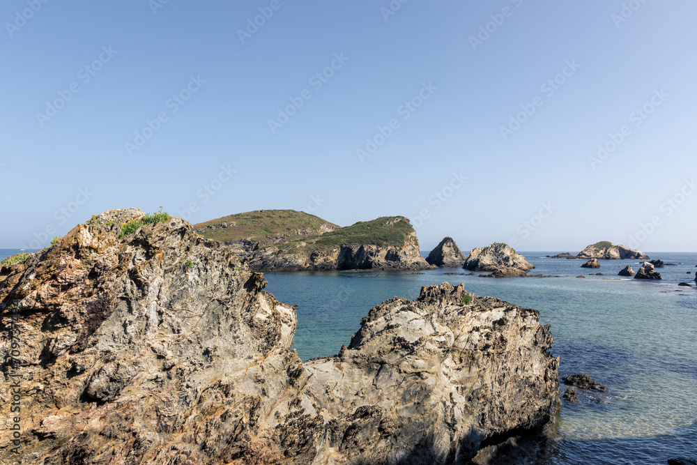 shows a scenic view of rocky formations, clear blue waters, and a hill under a clear sky