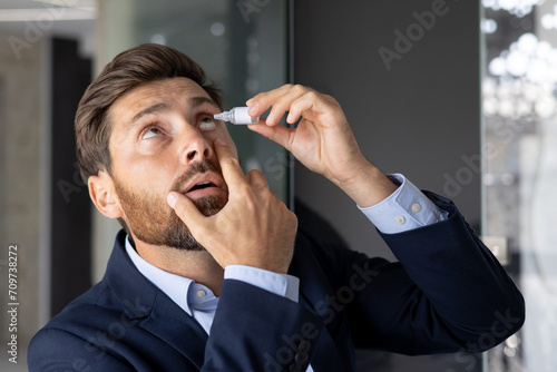 Close-up photo of a young businessman sitting in the office at a desk in a suit and instilling medicine into his eye, treating inflammation, pain, chronic fatigue photo
