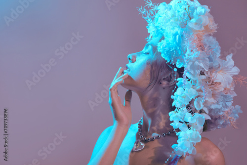 A sensual, gentle woman with a flower wreath on her head poses in blue lighting on a pink background