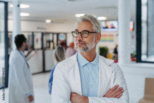 Portrait of confident mature doctor standing in Hospital corridor. Handsome doctor with gray hair wearing white coat, stethoscope around neck standing in modern private clinic, looking at camera. photo
