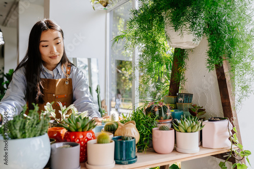 Small business, home decor concept. Lovely cute asian female floral store owner taking care, arranges flower pots, keeps track of plants at flower shop interior.
