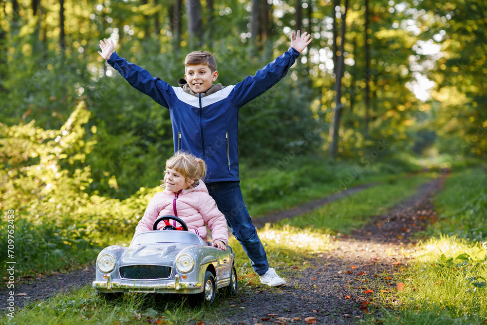 Two happy children playing with big old toy car in autumn forest, outdoors. Kid boy pushing and driving car with little toddler girl, cute sister inside. Laughing and smiling kids. Lovely family