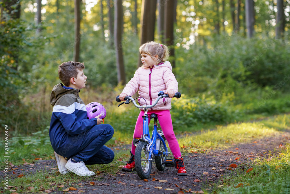 School kid boy, brother put on little preschool sister girl bike helmet on head. Brother teaching happy child cycling and having fun with learning bike. Active siblings family outdoors. Kids activity School kid boy, brother put on little preschool sister girl bike helmet on head. Brother teaching happy child cycling and having fun with learning bike. Active siblings family outdoors. Kids activity
