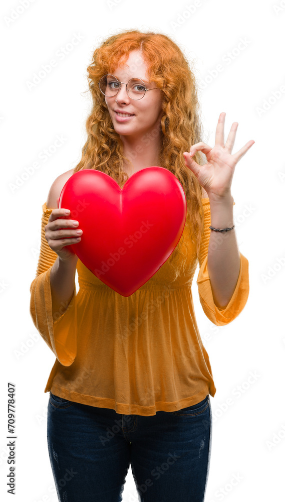 Young redhead woman in love holding red heart doing ok sign with fingers, excellent symbol Young redhead woman in love holding red heart doing ok sign with fingers, excellent symbol