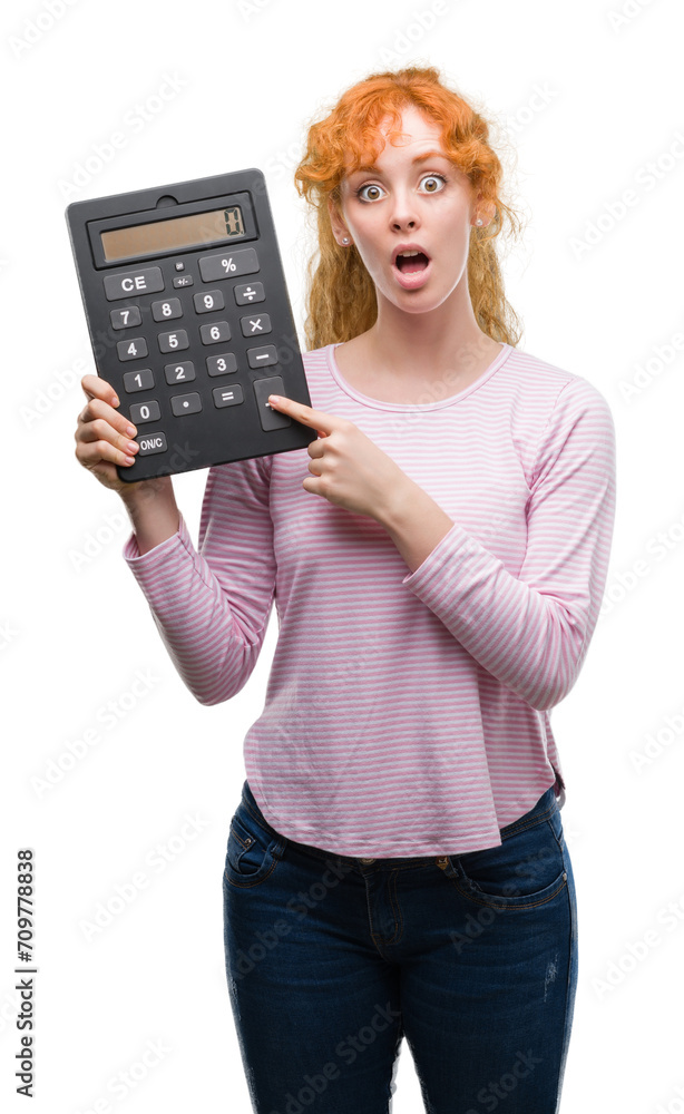 Young redhead woman holding big calculator scared in shock with a surprise face, afraid and excited with fear expression Young redhead woman holding big calculator scared in shock with a surprise face, afraid and excited with fear expression