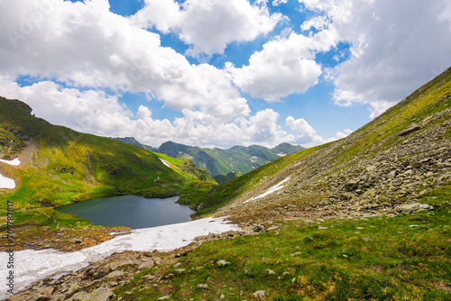 alpine landscape of fagaras mountains. capra lake of romania in summer. sunny weather with clouds on the blue sky. spots of snow and grass on the rocky hillside