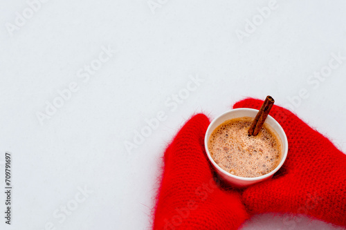 Red knitted mittens hold a cup of hot aromatic cocoa. Warmth, comfort, winter atmosphere, snow, nature. Winter hot drink with cinnamon and spices. Horizontal, free copy space.