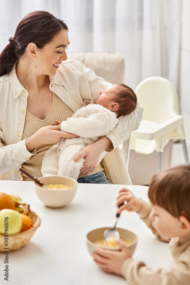 loving good looking mother having breakfast with her toddler son and holding her newborn baby loving good looking mother having breakfast with her toddler son and holding her newborn baby