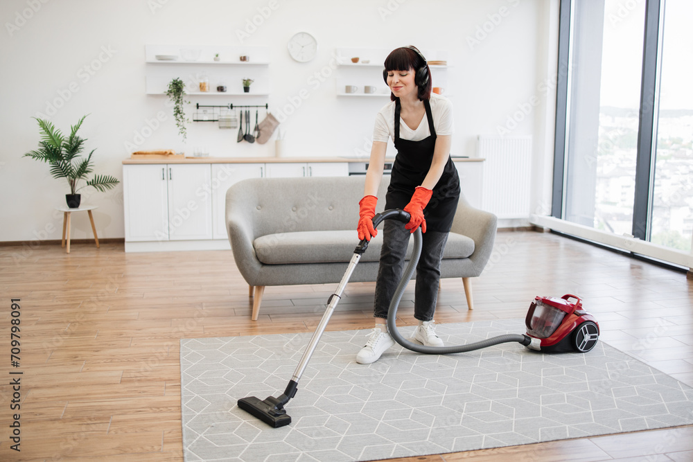 Positive young cleaners vacuuming rug of stylish modern studio kitchen. Cleaning service employee listens to music in headphones and dances while cleaning bright and spacious apartment.