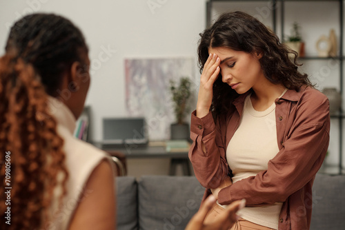 Offended and upset brunette girl touching forehead while standing in front of her irritated girlfriend making accuses during quarrel photo