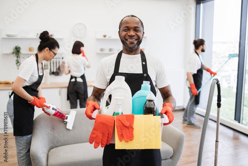 Portrait of young African American man professional cleaning worker holding a bucket for washing with detergents on bright kitchen studio background, copy space.