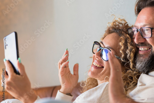 Happy Young Adult Couple Making Video Call On Phone - cheerful people speak and listen friends online with cellular from home sitting on the sofa - holidays remote celebration concept