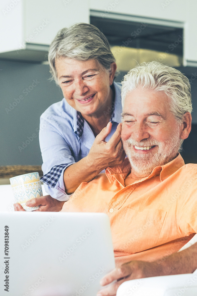 Elderly real lifestyle people using computer at home. Leisure indoor activity with man and woman enjoying laptop and internet connection sitting on the sofa with kitchen in background Elderly real lifestyle people using computer at home. Leisure indoor activity with man and woman enjoying laptop and internet connection sitting on the sofa with kitchen in background