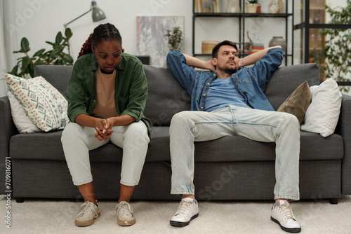 Young upset African American woman sitting on couch next to her indifferent husband looking aside during quarrel