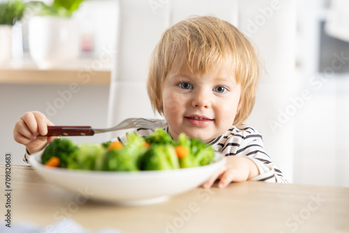 Cute little girl is eating broccoli and carrot vegetables with a fork