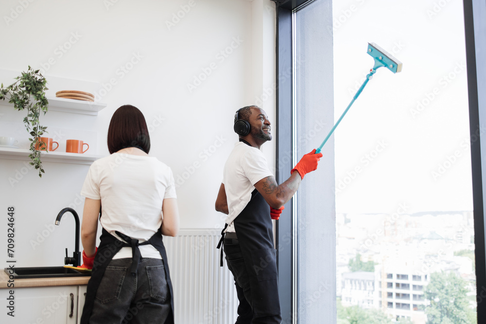 Multiethnic employees of company cleaning in bright modern loft kitchen with panoramic windows. Young African washing windows with mop, listening to music in headphones and dancing. Multiethnic employees of company cleaning in bright modern loft kitchen with panoramic windows. Young African washing windows with mop, listening to music in headphones and dancing.