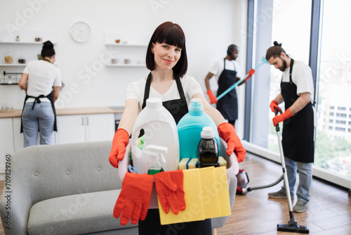 Portrait of young Caucasian woman professional cleaning worker holding a bucket for washing with detergents on bright kitchen studio background, copy space.