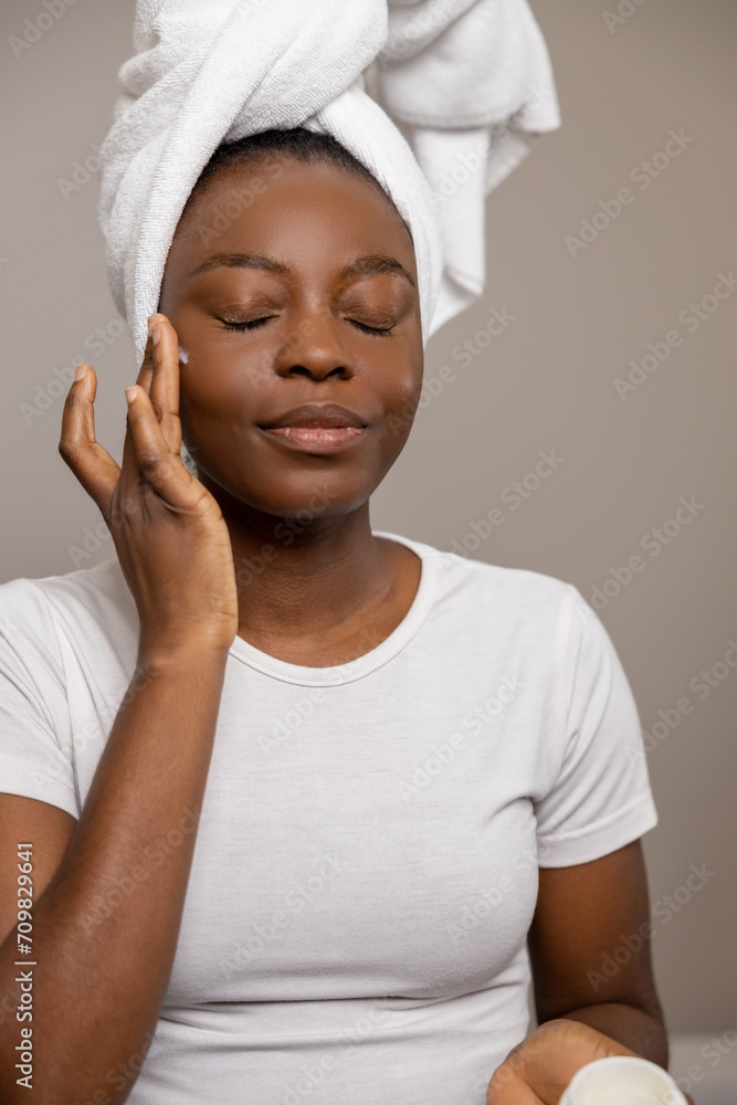 Satisfied woman applying daily care cream closing her eyes standing with towel on head Satisfied woman applying daily care cream closing her eyes standing with towel on head