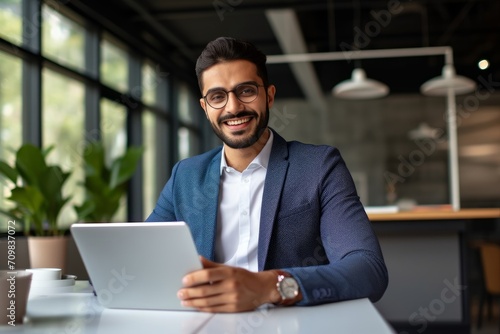 Happy Indian business man manager analyst using tablet computer, businessman employee or executive looking at camera working on tab device with financial data ai solutions at office desk.Generative AI